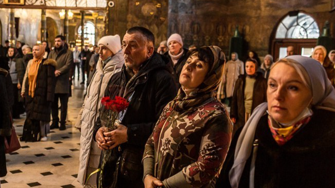 A group of people stand closely together inside a church, some wearing headscarves. One man holds red flowers, and everyone appears solemn, with warm light illuminating the scene.