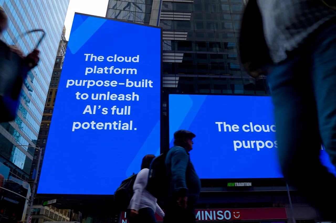 People walk past two large digital billboards in a city displaying the message: “The cloud platform purpose-built to unleash AI’s full potential.” The scene is set in a busy urban area with tall buildings.