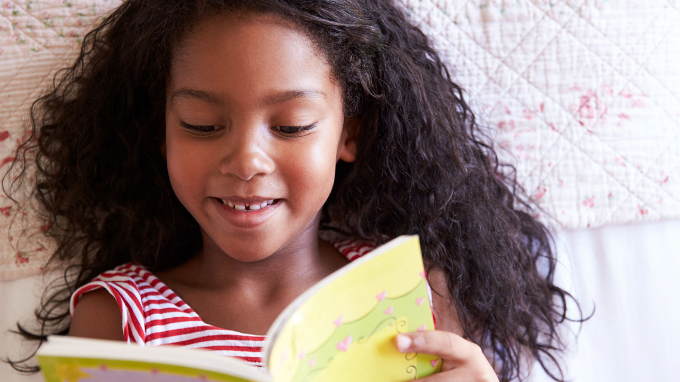 A young girl with long curly hair, wearing a red and white striped top, smiles while reading a yellow book. She is sitting on a bed with a white patterned quilt in the background.