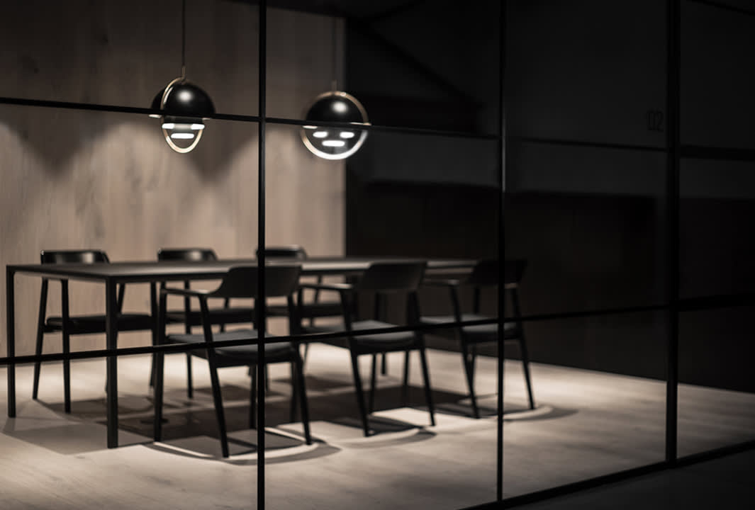 A modern, dimly lit meeting room features a black table surrounded by six black chairs. Two spherical pendant lights hang above the table, casting focused light, while the room is enclosed by glass walls.