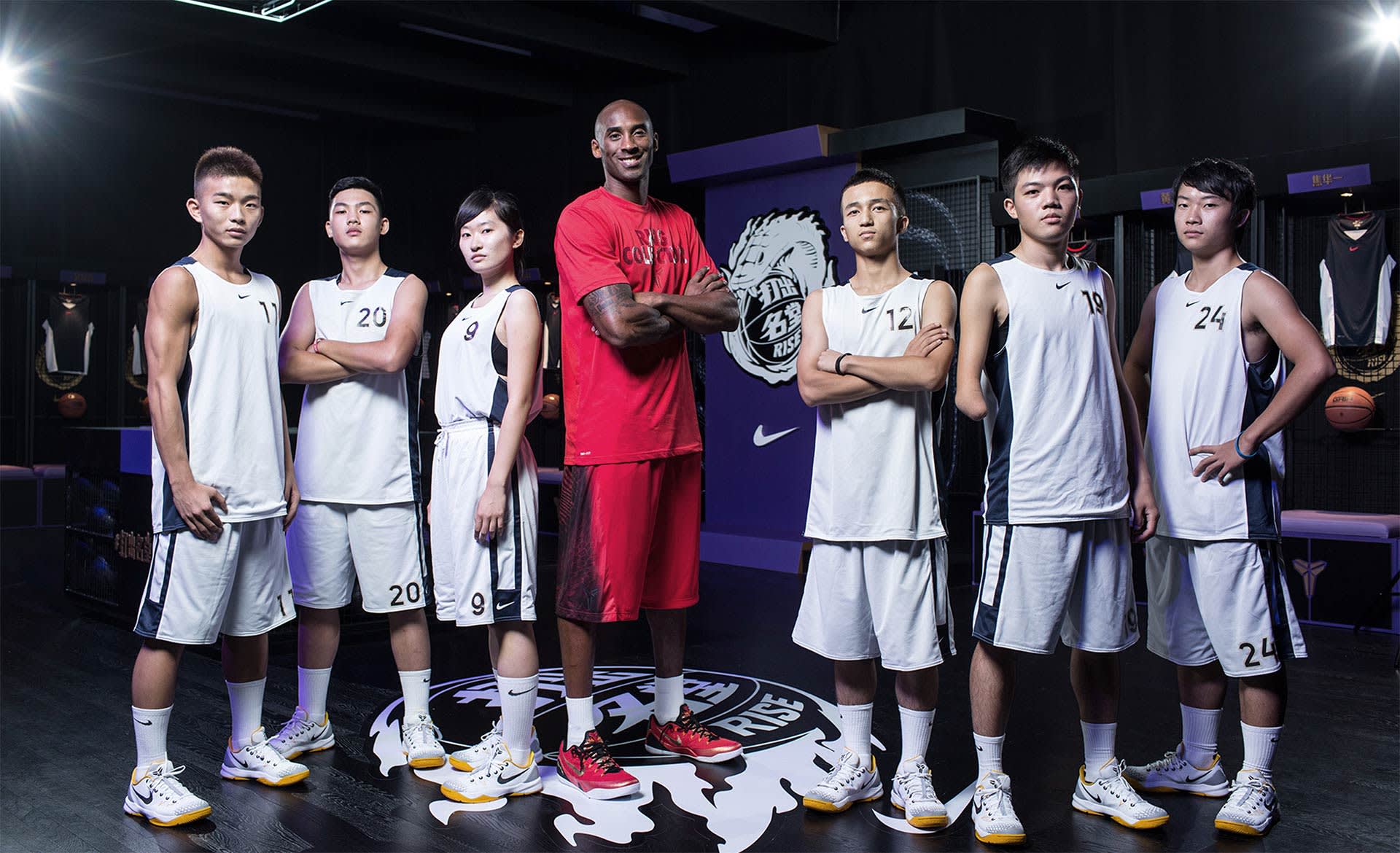 A group of six young basketball players in white uniforms stand alongside a tall man in a red shirt and shorts inside a dimly lit gym, all looking at the camera with confident expressions. Sports equipment and jerseys are visible in the background.