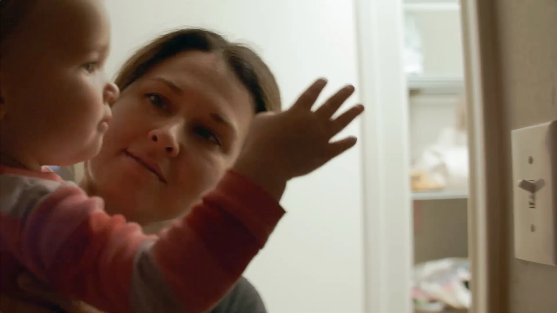 A woman smiles gently while holding a young child who raises their hand near a light switch in a softly lit room. Shelves with various items are visible in the background.