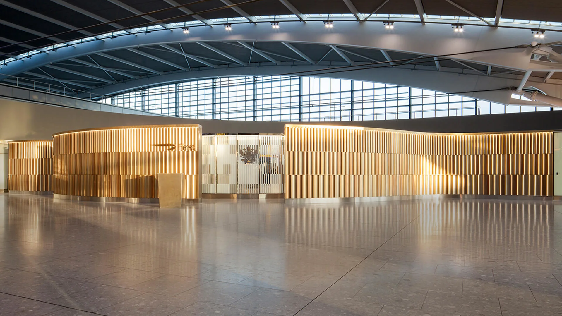 A modern airport lounge features a sleek, curved wall with vertical golden slats, large glass windows above, and polished stone flooring, illuminated by natural and artificial light.