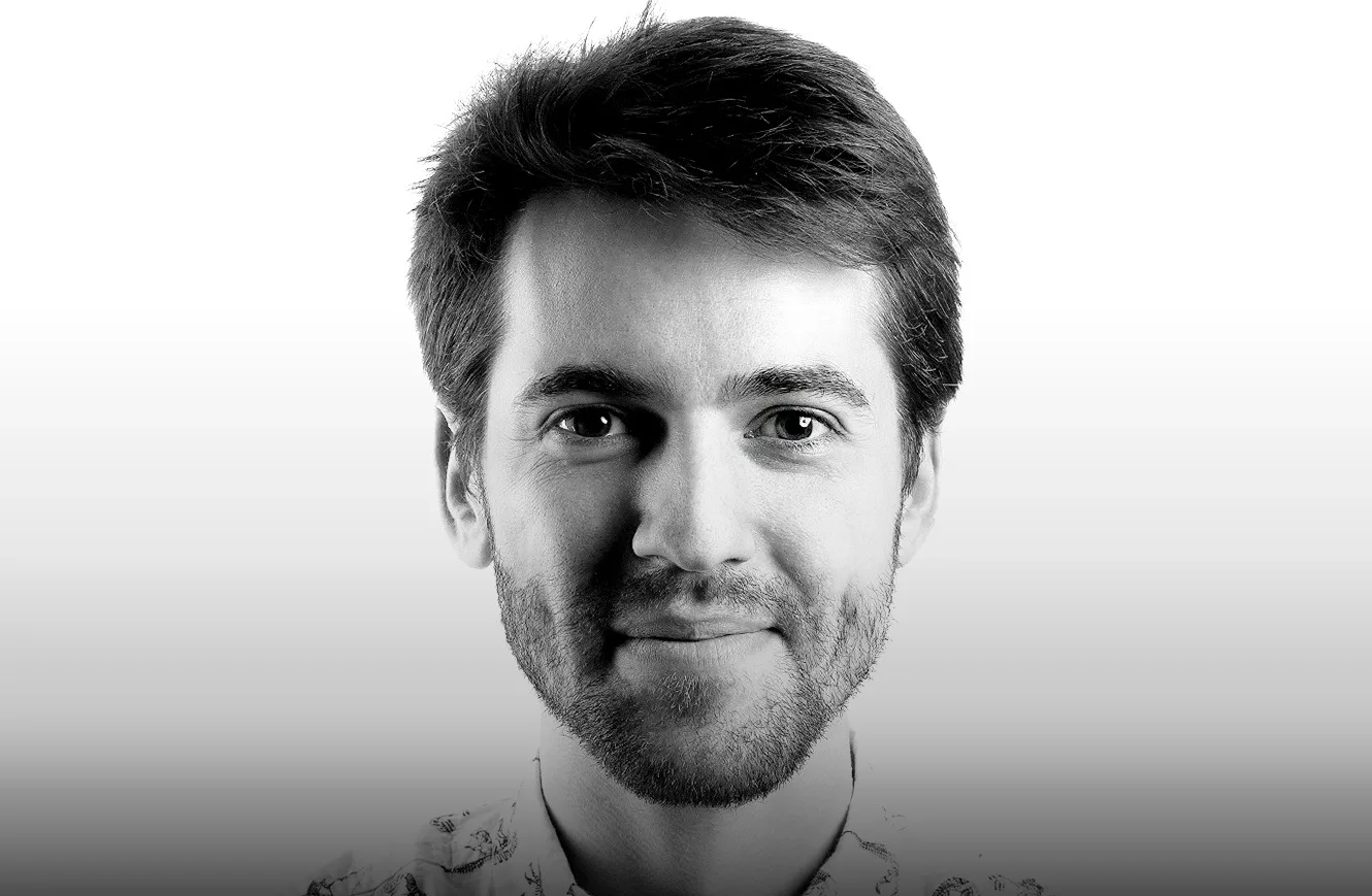 Black and white portrait of a young man with short hair and a beard, smiling softly, looking directly at the camera against a plain white background.