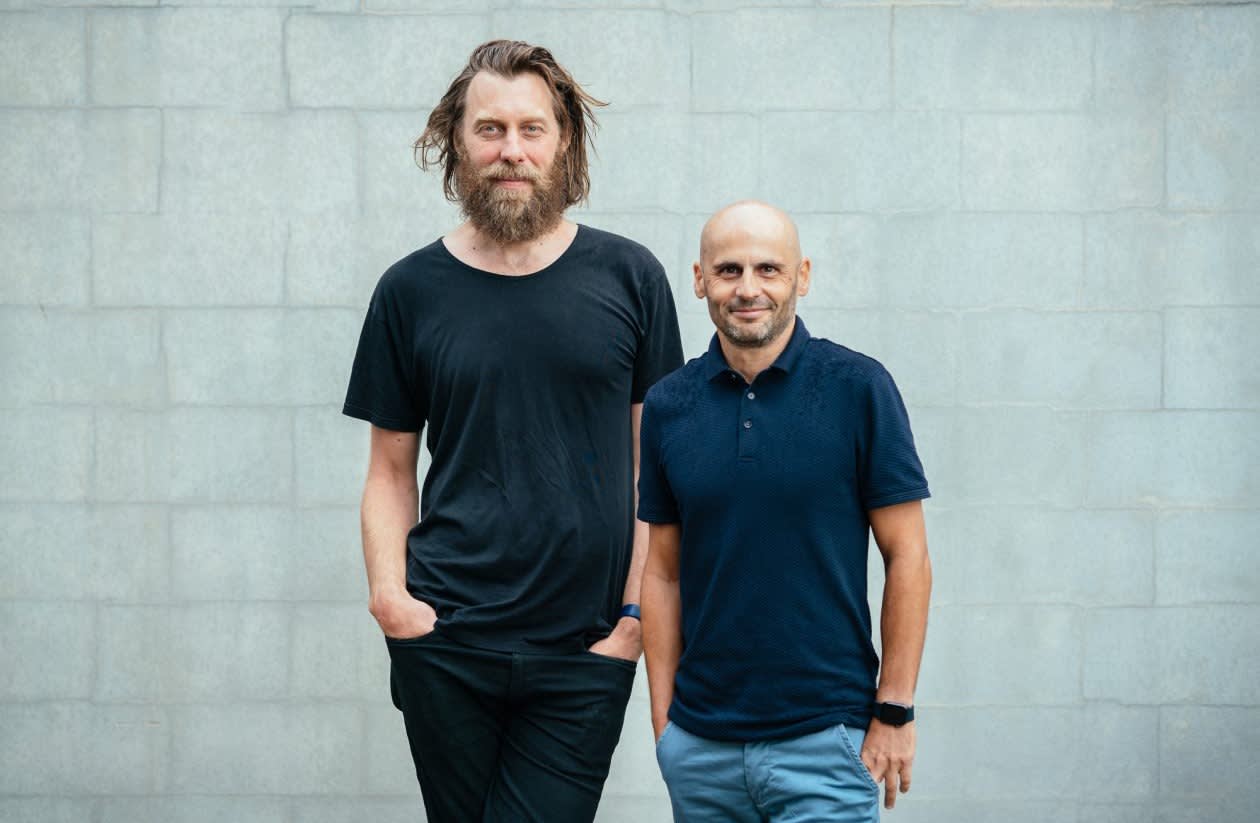 Two men standing side by side in front of a gray brick wall. The taller man on the left has long hair, a beard, and wears a black t-shirt and pants. The shorter man on the right is bald and wears a navy polo shirt and blue pants.