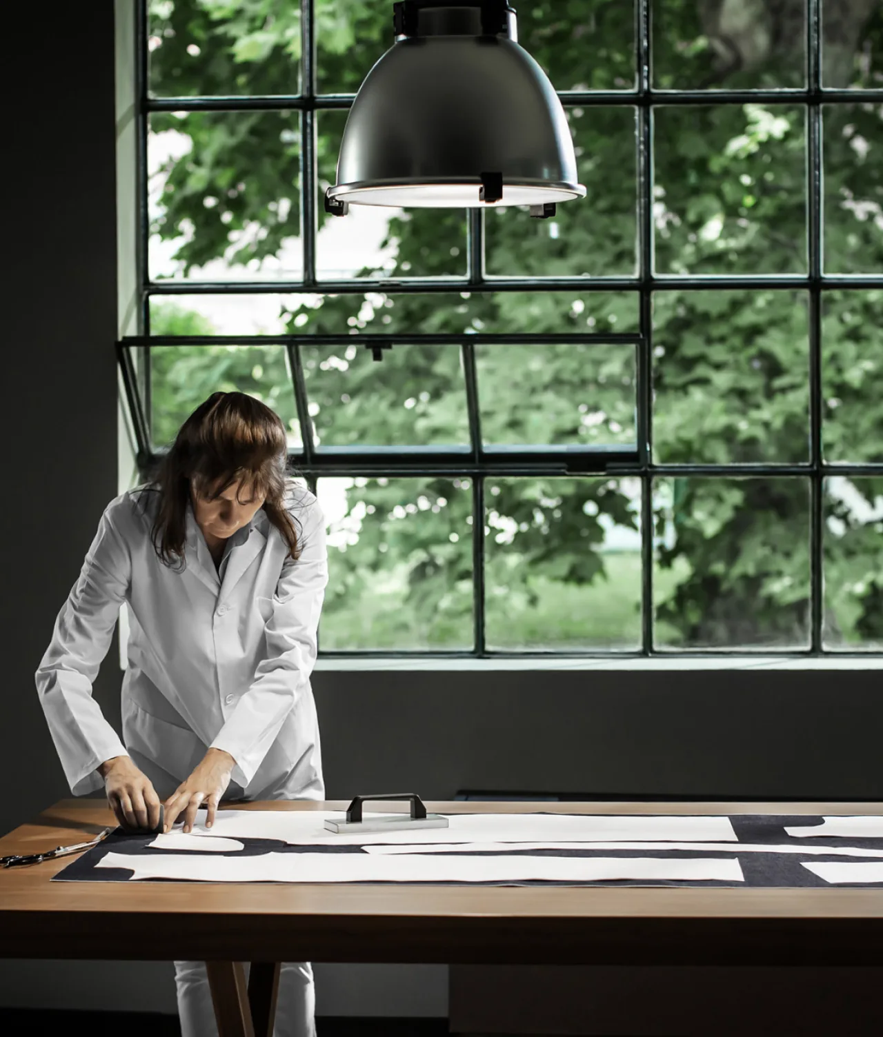 A person in a white coat works on a large pattern or fabric on a wooden table in front of a tall window with green trees outside, illuminated by a large overhead lamp.