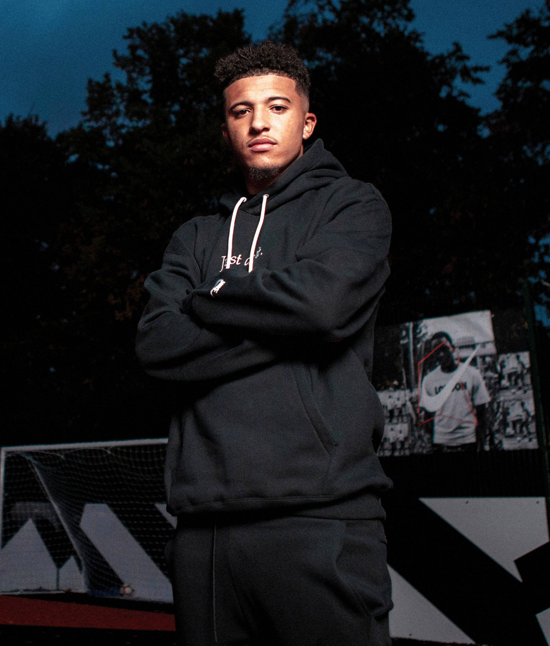 A young man wearing a black hoodie and pants stands with his arms crossed in front of a soccer goal and a mural, with trees and a darkening sky in the background.