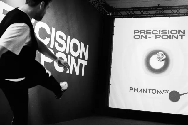 A person kicks a soccer ball toward a digital screen displaying the words PRECISION ON POINT and PHANTOM with a target graphic. The scene appears to be part of a sports exhibit or promotional event.
