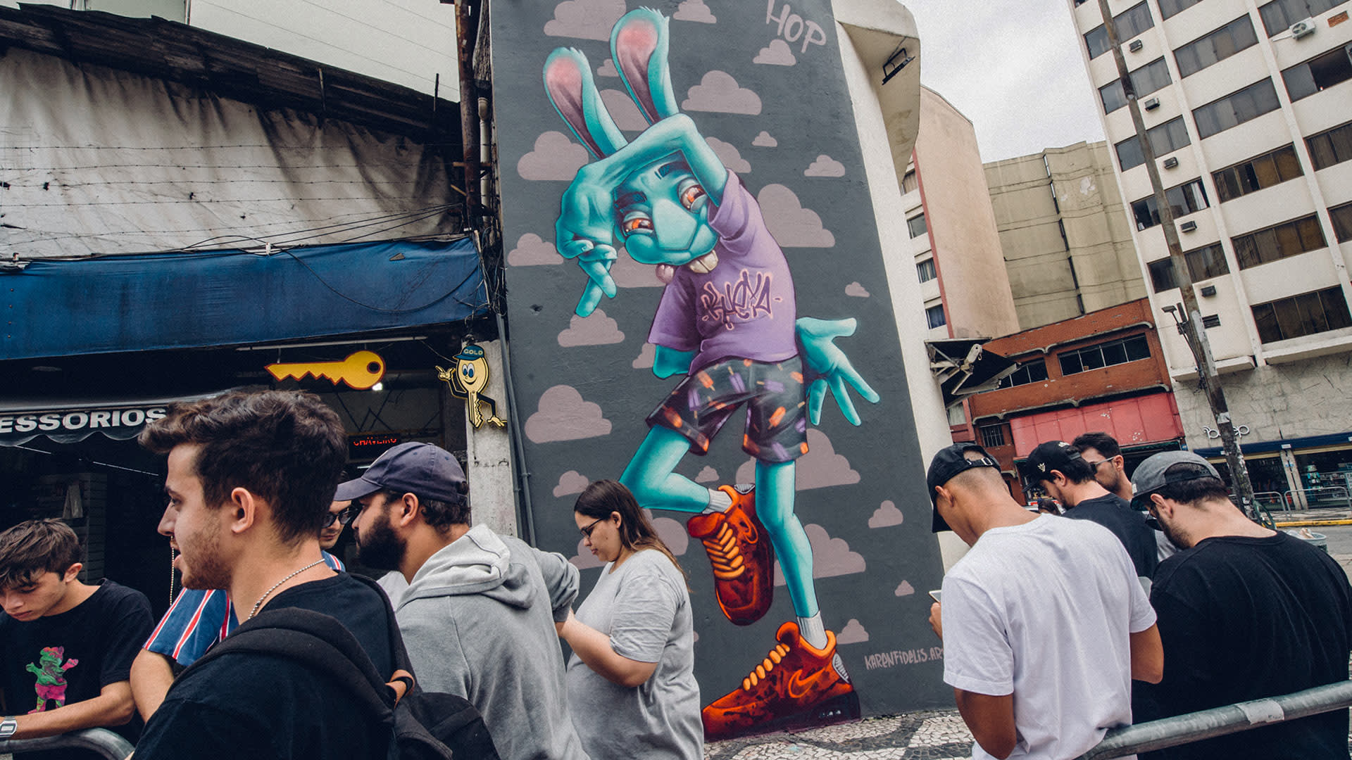 A group of people stand in line next to a tall mural of a blue, humanoid rabbit wearing streetwear and orange sneakers, painted on a city building wall. The sky is overcast and buildings surround the scene.