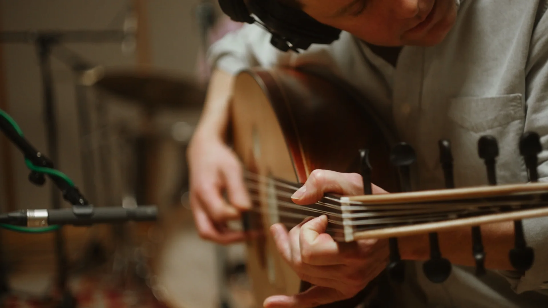 A person wearing headphones plays an oud, a traditional string instrument, in a recording studio. A microphone and drums are visible in the background. The focus is on the musicians hands plucking the strings.
