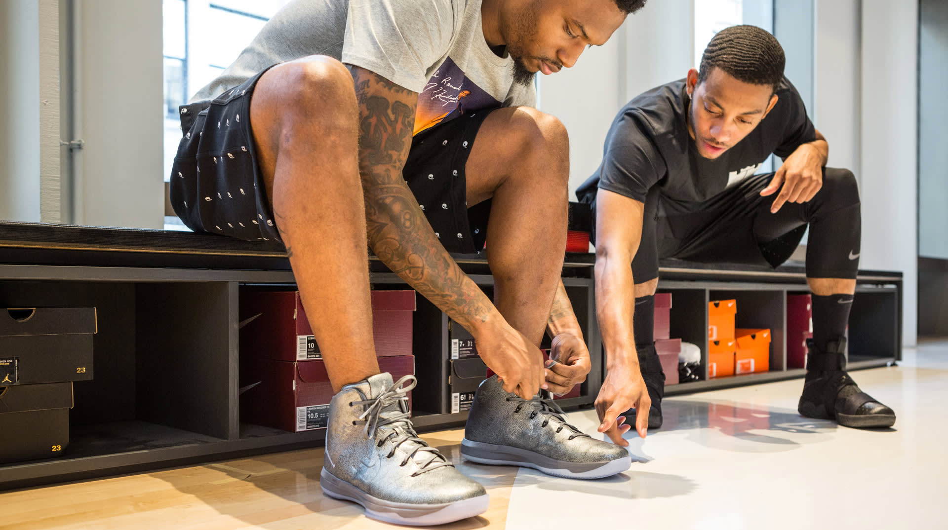 Two men sit in a shoe store; one is tying the laces of shiny silver basketball shoes while the other points at the shoe, surrounded by shelves with colorful shoeboxes.