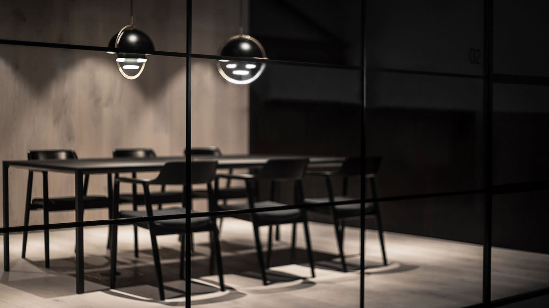 A modern conference room with a black table, six black chairs, and two round pendant lights hanging above, seen through glass walls. The room has minimalistic decor and a neutral color palette.