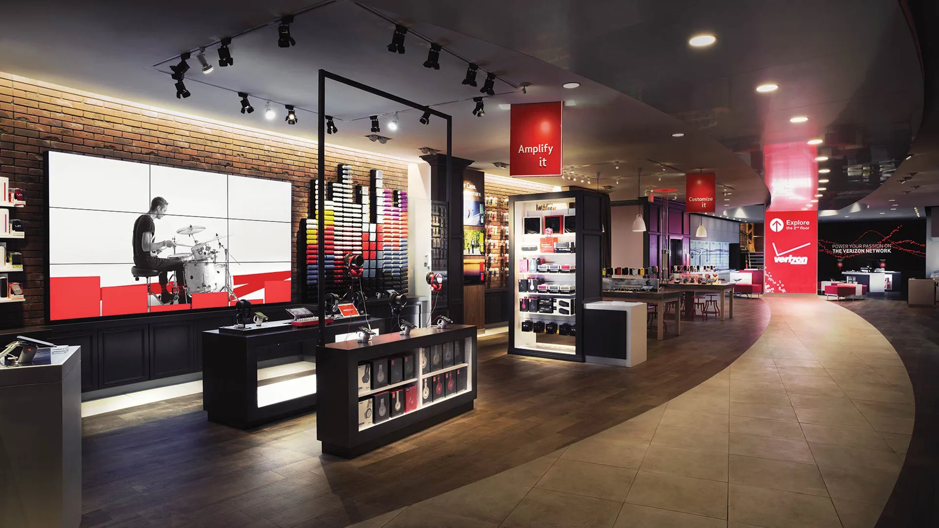 A modern Verizon store interior with digital displays, shelves of electronics and accessories, a large screen showing a drummer, and bright lighting highlighting various tech products.