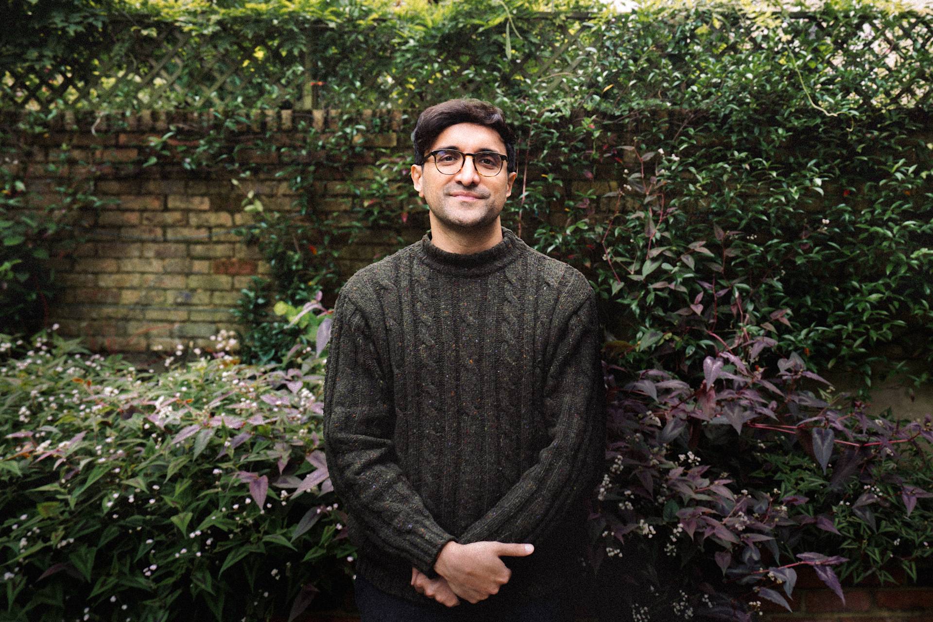 A person with short dark hair and glasses stands outdoors in front of a brick wall and dense green foliage, wearing a dark knit sweater and smiling gently at the camera.