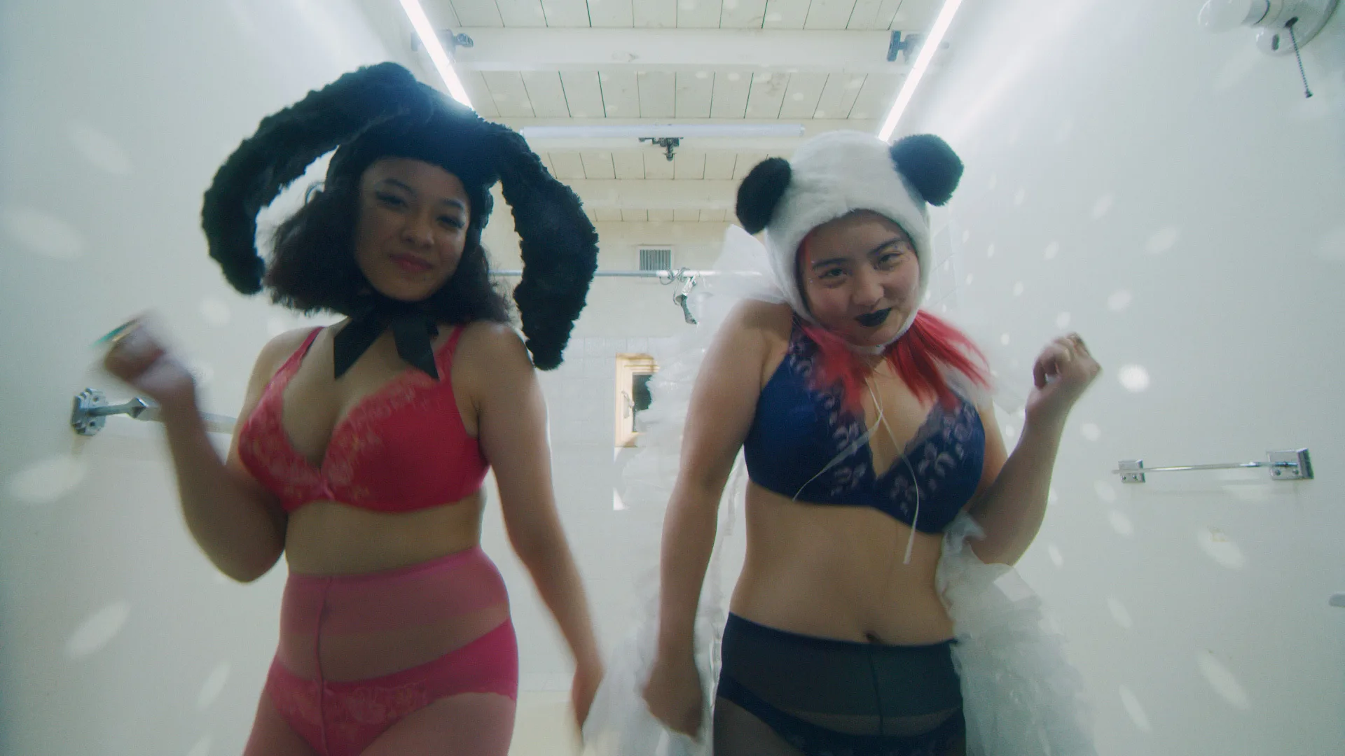 Two people wearing animal ear headpieces and colorful lingerie, smiling and dancing in a brightly lit, white-tiled room with lights reflecting off the walls.