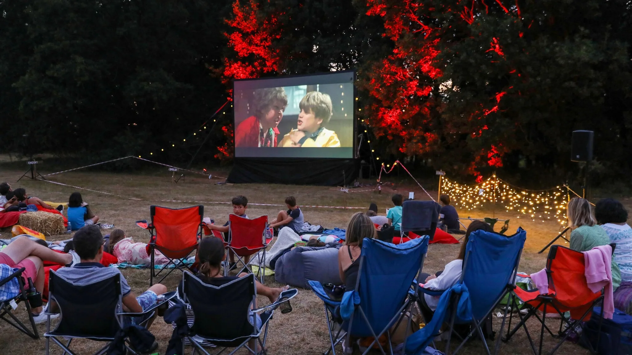 People sit on lawn chairs and blankets in a grassy area at night, watching a movie on a large outdoor screen. Trees in the background are lit with red and yellow lights, and string lights add a festive atmosphere.
