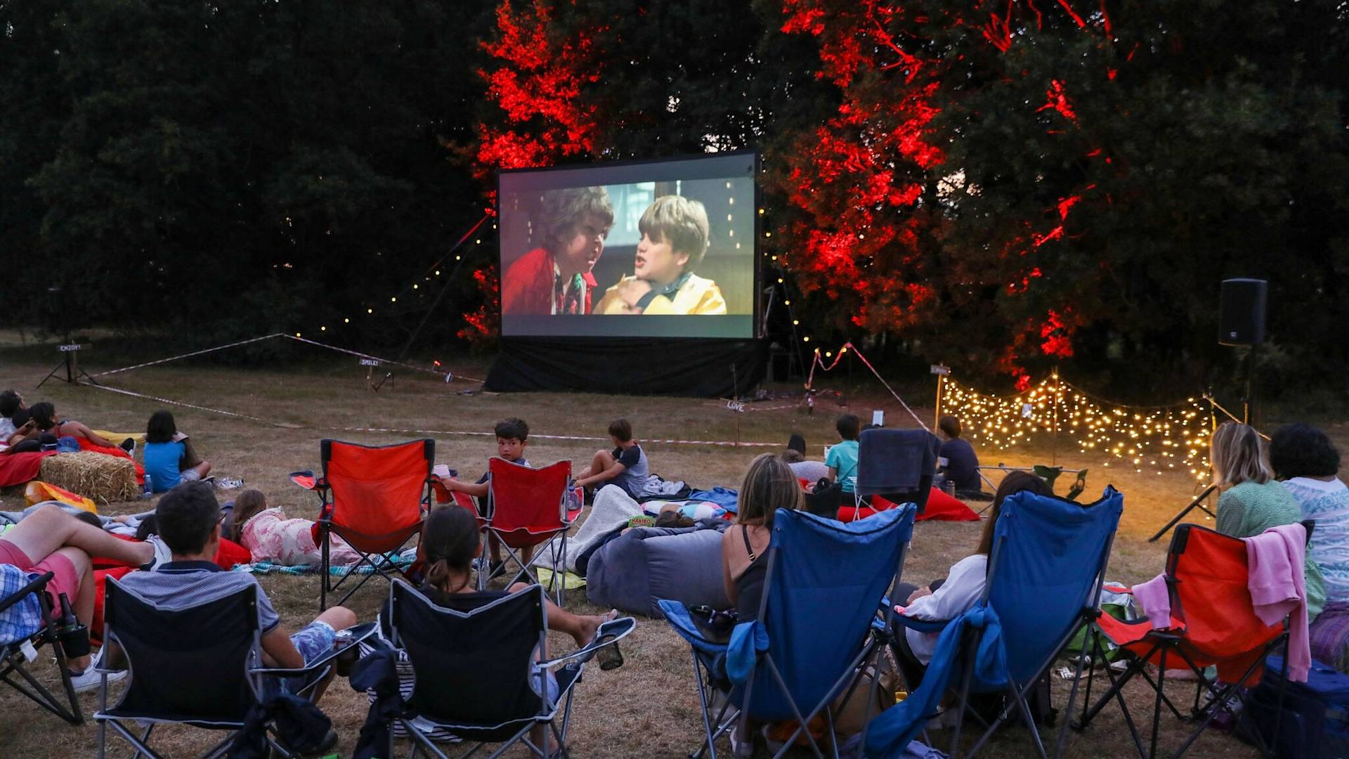 People sit on lawn chairs and blankets in a grassy area at night, watching a movie on a large outdoor screen. Trees in the background are lit with red and yellow lights, and string lights add a festive atmosphere.