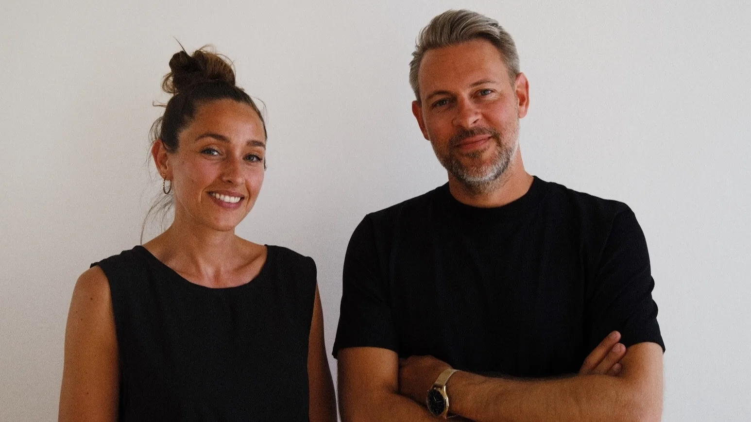 A woman and a man stand side by side against a plain white wall, both smiling. The woman has her hair in a bun and wears a sleeveless black top; the man has short gray hair, a beard, and wears a black t-shirt with his arms crossed.