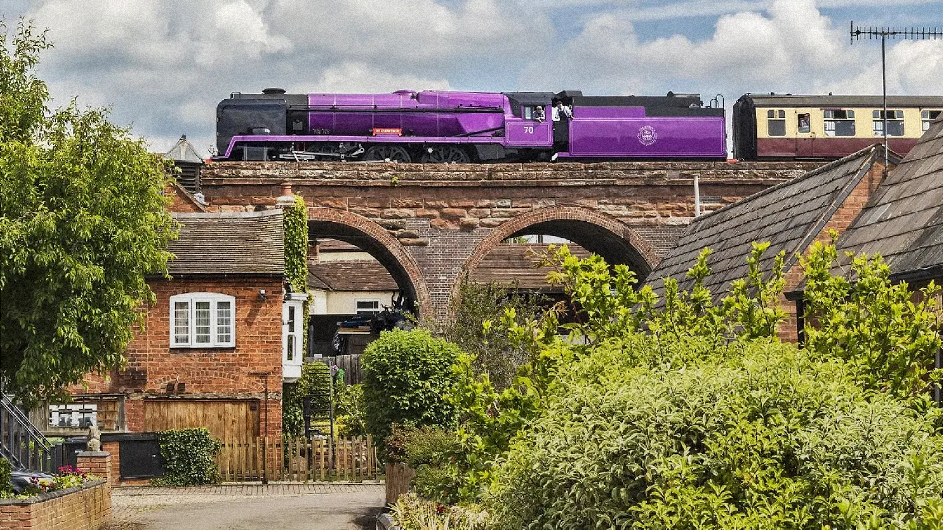 A purple steam train crosses a brick viaduct above quaint houses and lush green gardens on a sunny day with a partly cloudy sky.