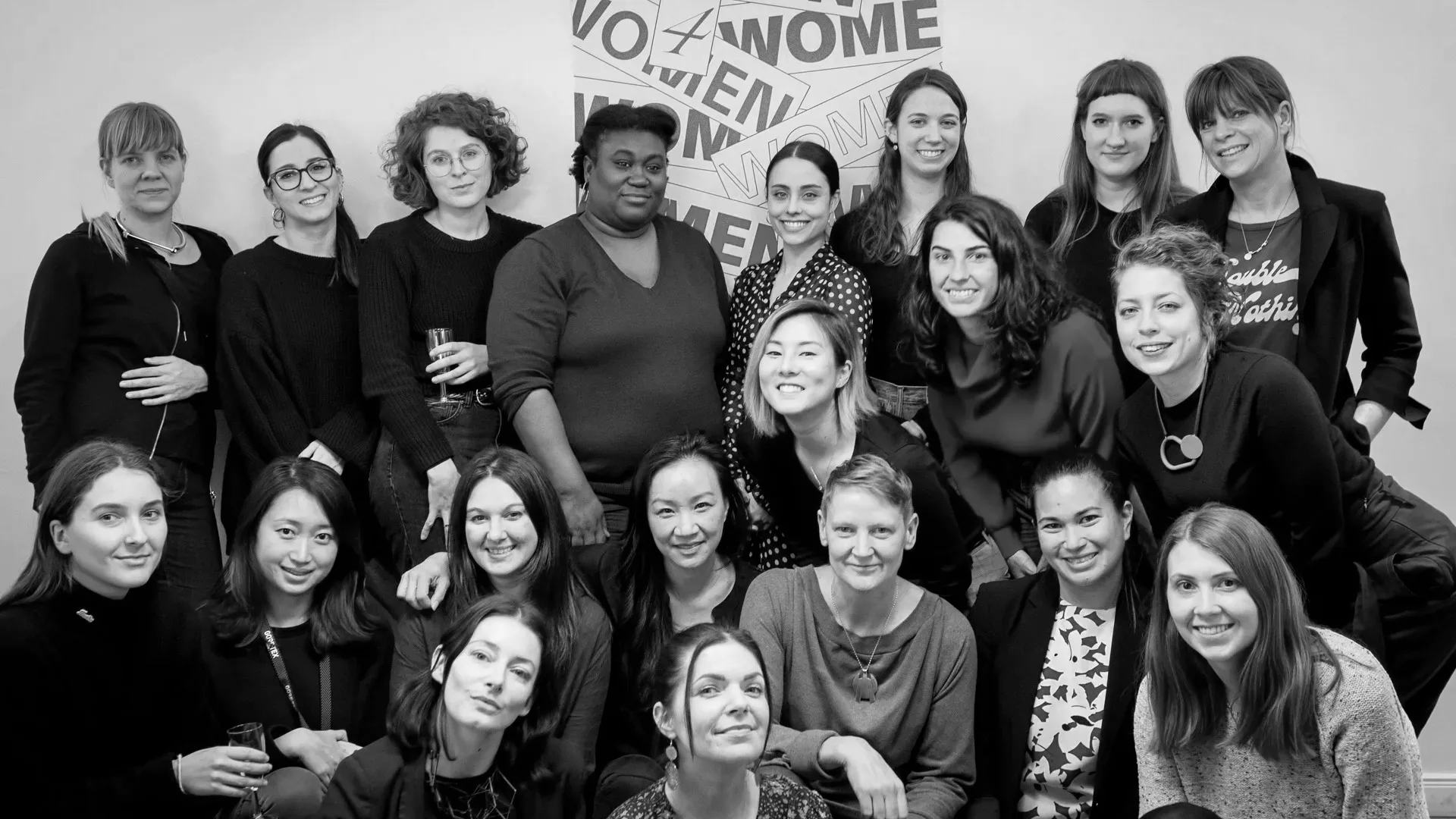 A group of twenty women pose together and smile for a photo in front of a wall with a Women 4 Women poster. Some women are seated, others are standing, and the image is in black and white.