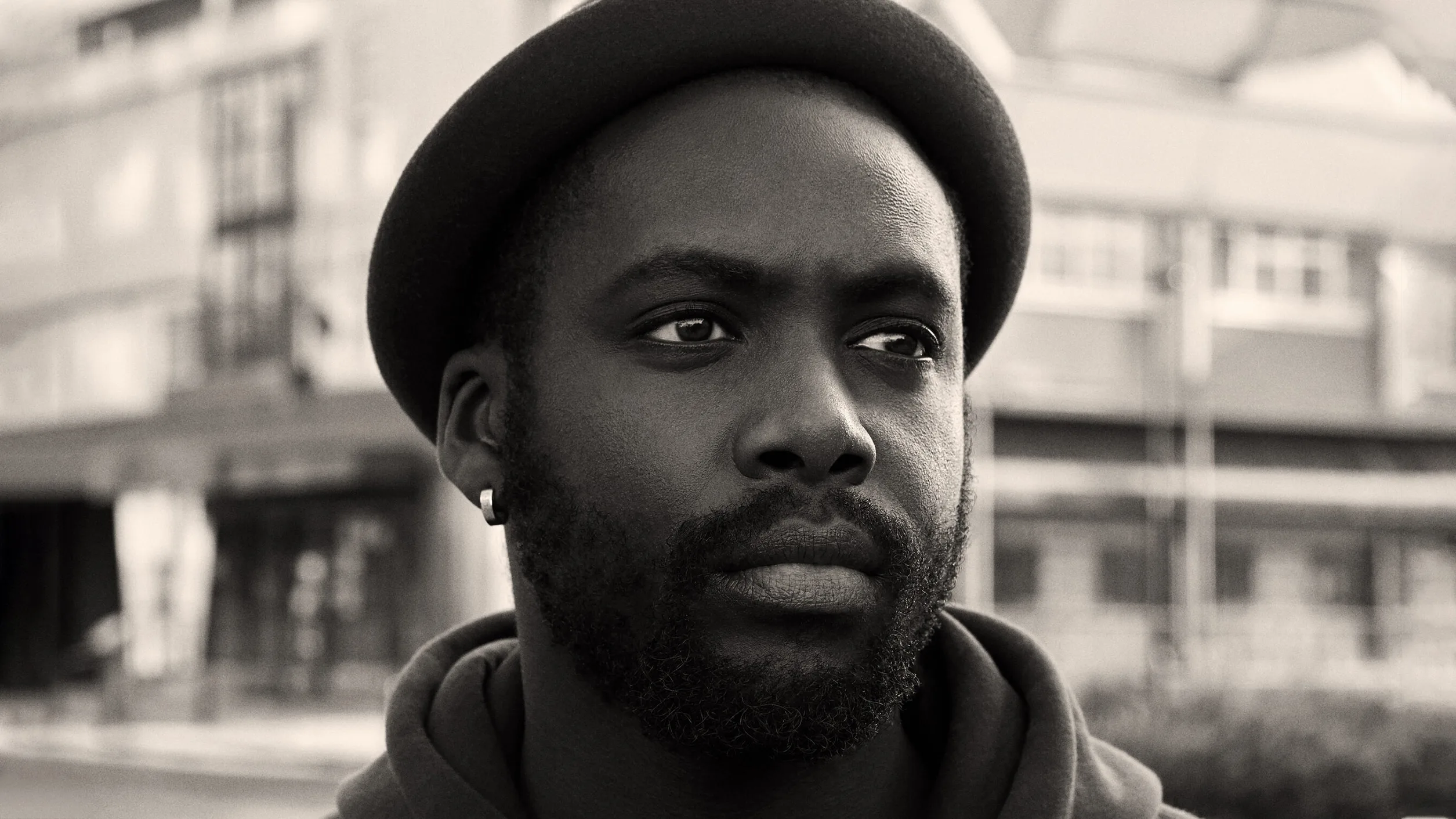 A man wearing a hat and hoodie looks pensively into the distance outdoors. The image is in black and white, with blurred urban buildings in the background.