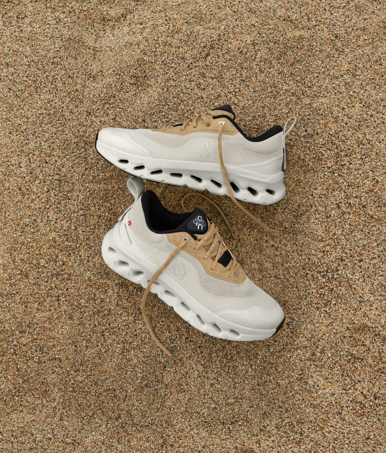 A pair of beige and tan athletic sneakers with white soles are placed on sandy ground, with the shoelaces loosely arranged.