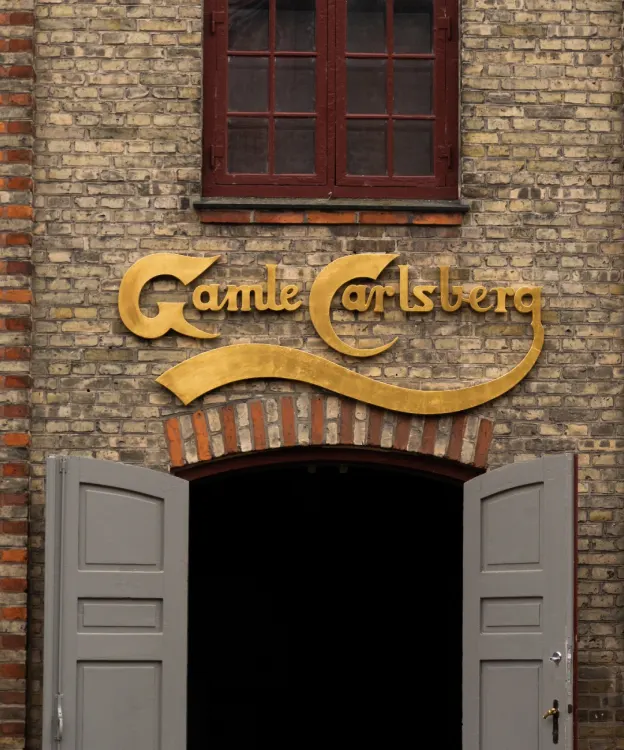 A brick building facade with a gold sign reading Gamle Carlsberg above an arched doorway with double grey doors, and a window with red framing above the sign.