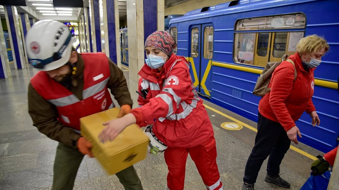 Red Cross workers in red uniforms and masks distribute a box of supplies in a subway station next to a blue train, while another person walks nearby.