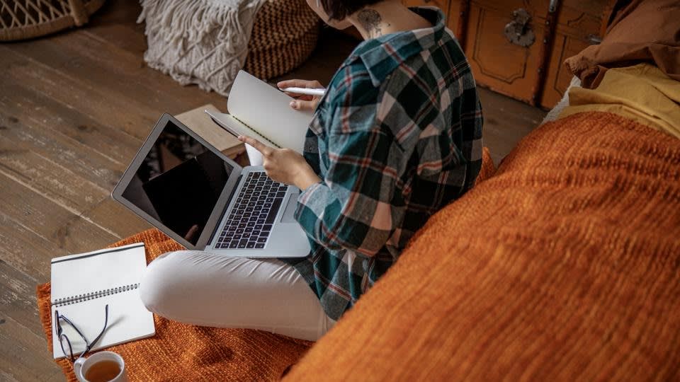 Person sitting on an orange blanket with a laptop on their lap, looking at an open notebook. A coffee cup, glasses, and another notebook are on the floor beside them. The scene suggests remote work or study at home.