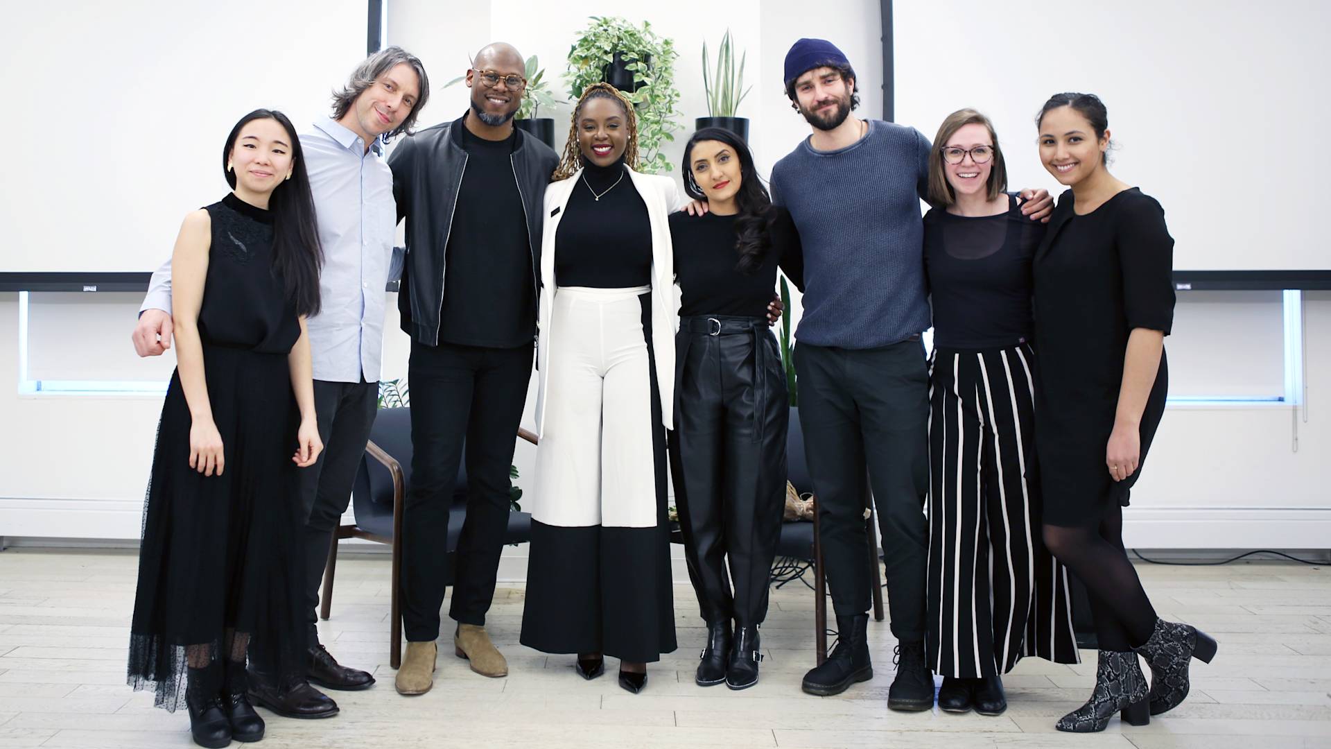 Eight people stand side by side indoors, smiling at the camera. They are dressed in black and white clothing and are posing in a modern room with plants and large windows in the background.