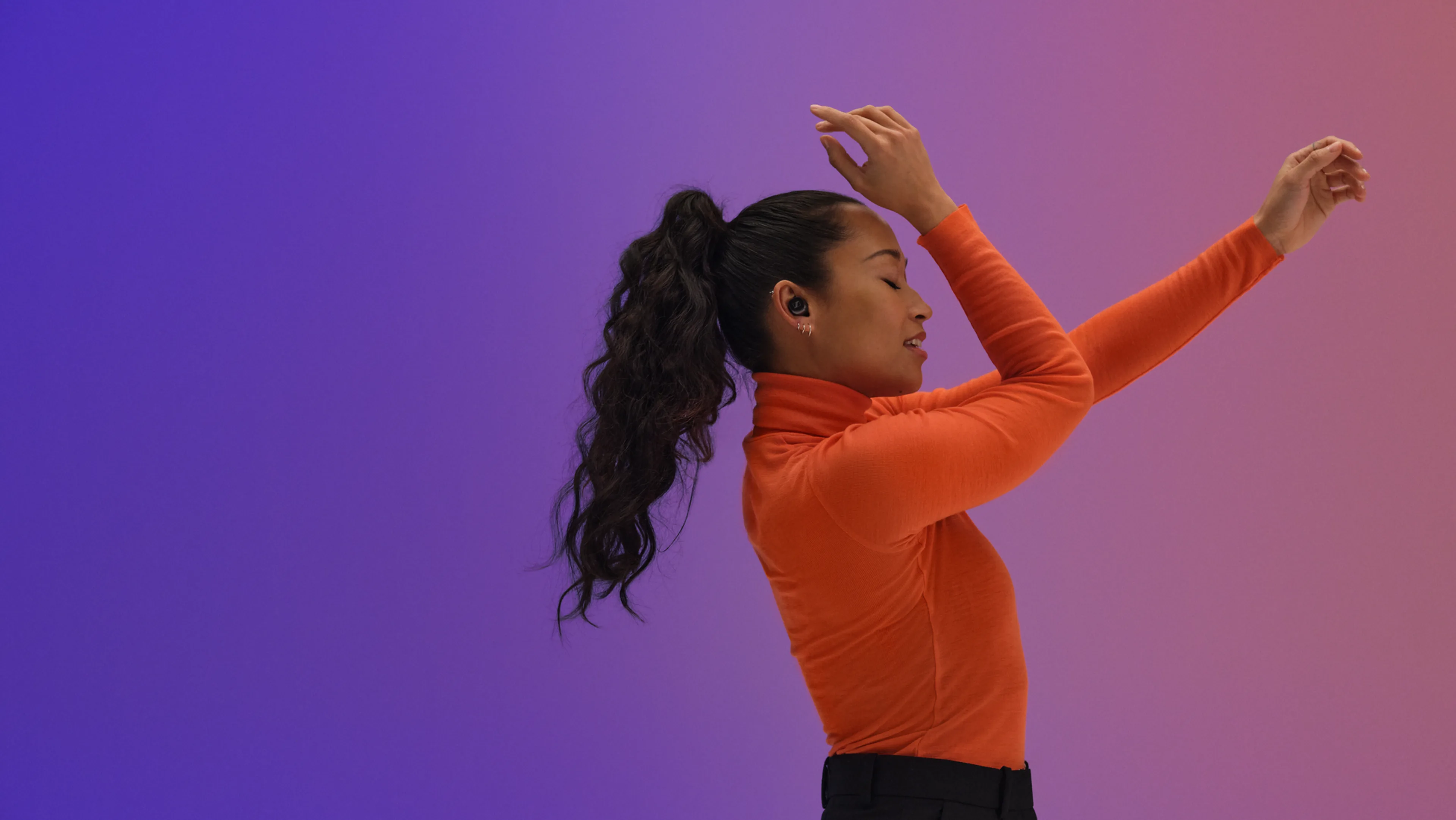 A woman with long curly hair in a ponytail wears an orange turtleneck and wireless earbuds, dancing with her arms raised against a gradient purple and pink background.