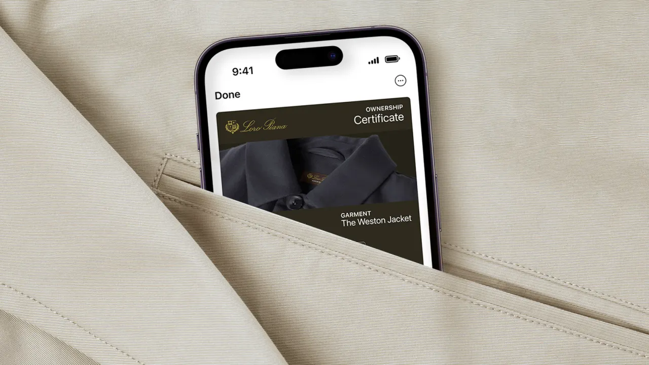A smartphone displaying a digital ownership certificate for The Weston Jacket is partially tucked into the pocket of a beige jacket. The screen shows a garment image and certificate details.