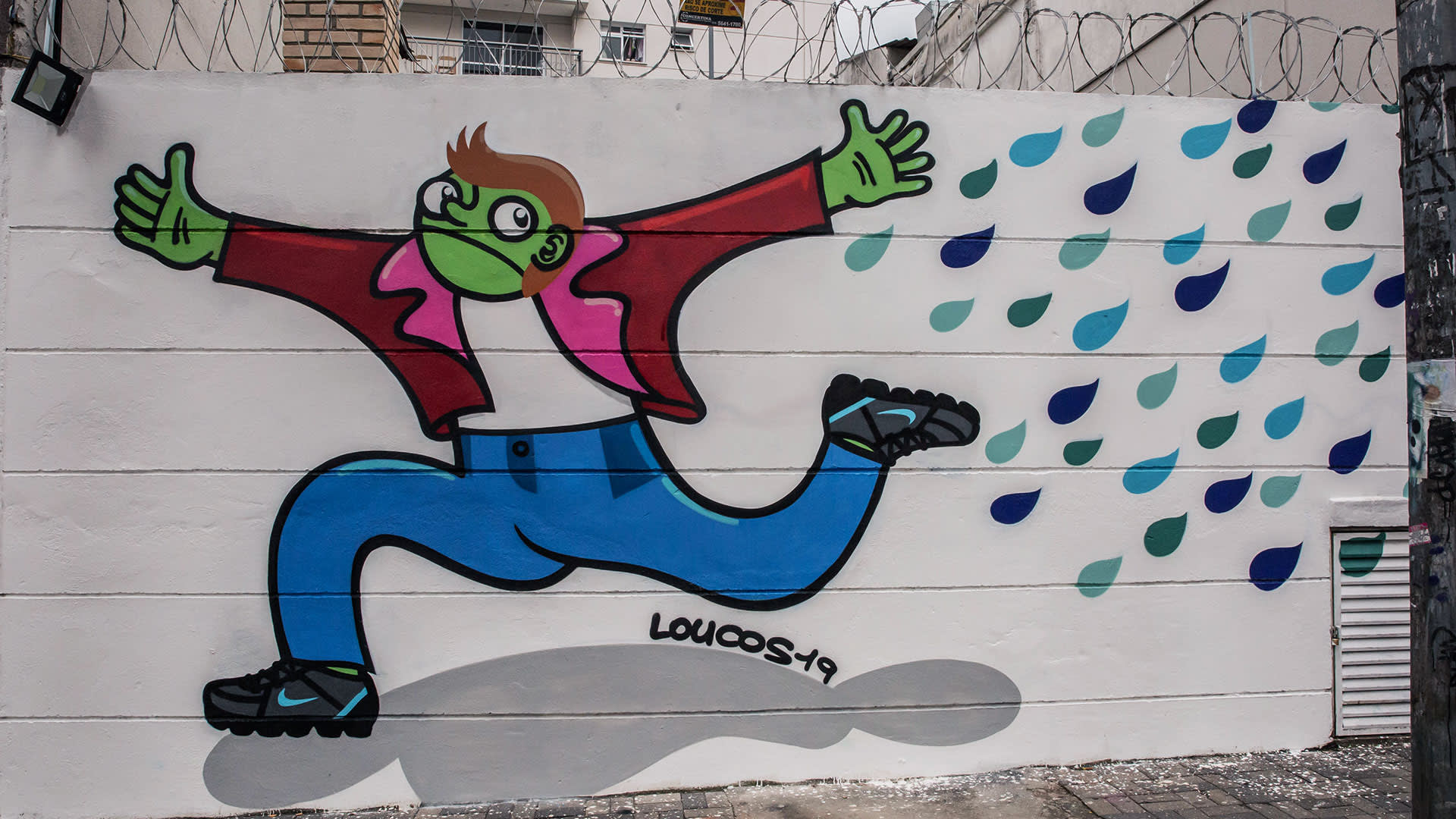 Street art mural of a cartoon figure with green skin, red jacket, blue pants, and a face mask, running joyfully with blue and teal raindrops in the background. The signature LOUCOS49 is visible at the bottom.