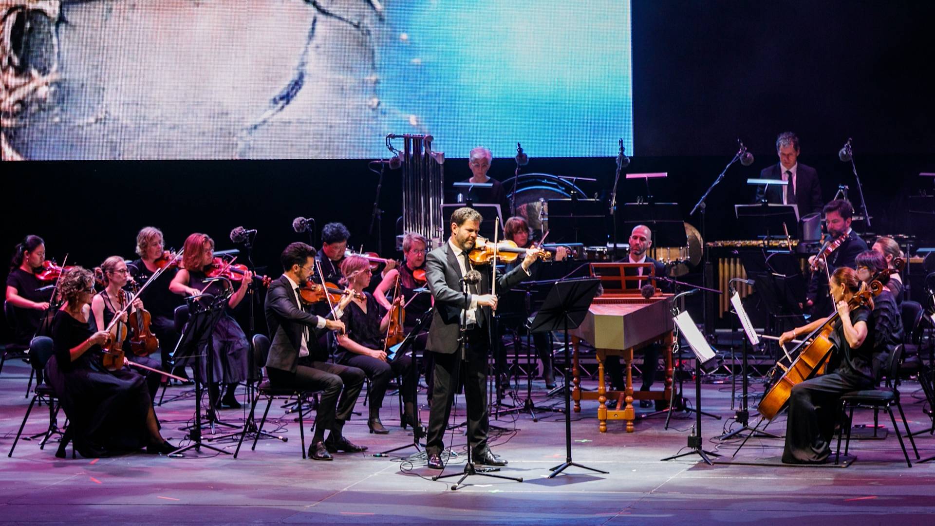 A symphony orchestra performs on stage, with musicians playing strings and percussion. A violinist stands at the front as soloist, while a large screen displays abstract visuals behind them. Music stands and microphones are visible.