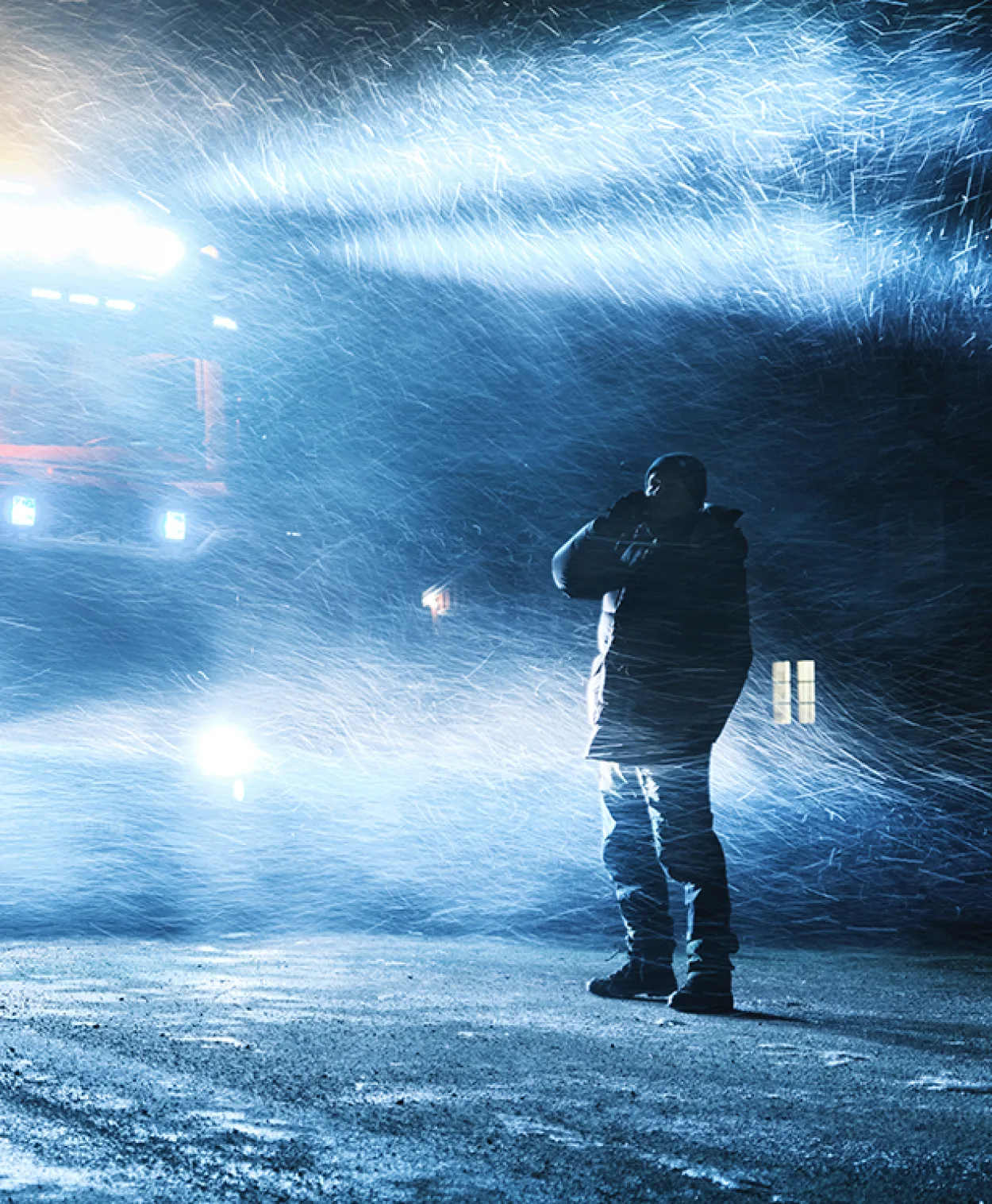 A person stands outside in a heavy snowstorm at night, illuminated by bright headlights shining through swirling snow. The scene is cold and dramatic, with snow blowing fiercely around them.