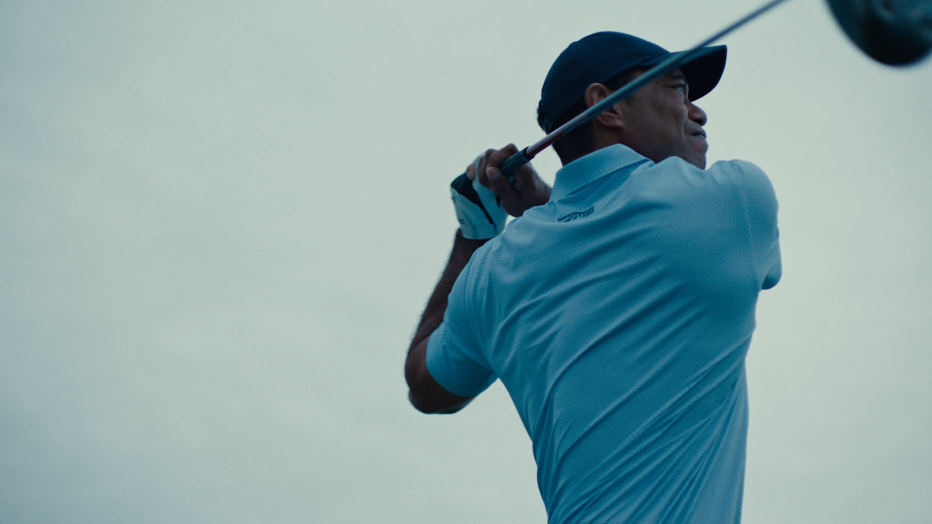Tiger Woods in a light blue shirt and dark cap swings a golf club against a bright, overcast sky, captured from a low angle behind him.