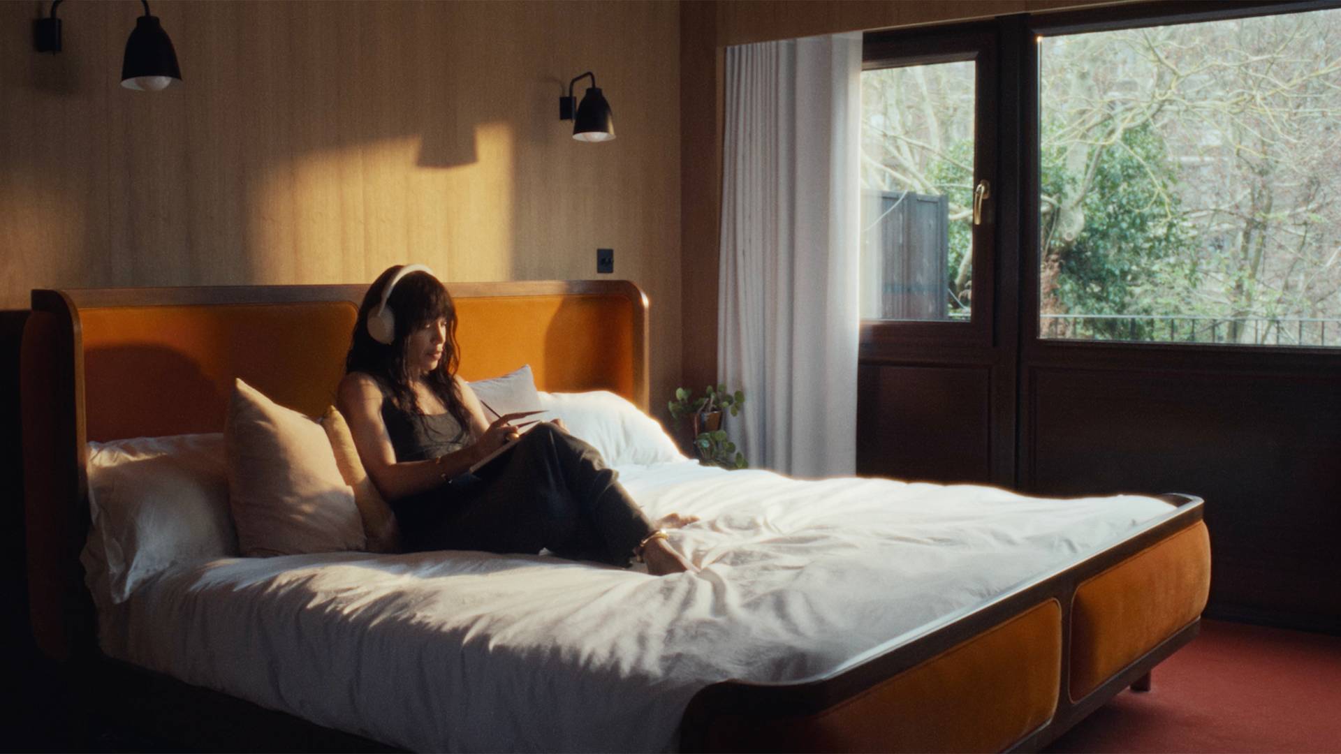Loreen wearing headphones sits on a large, neatly made bed in a softly lit room, reading or using a tablet. Sunlight streams through a window, highlighting greenery outside.