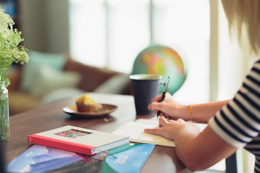 A person writes in a notebook at a table with a book, a muffin on a plate, a cup, and a globe in the background. A vase of flowers is also visible on the left.