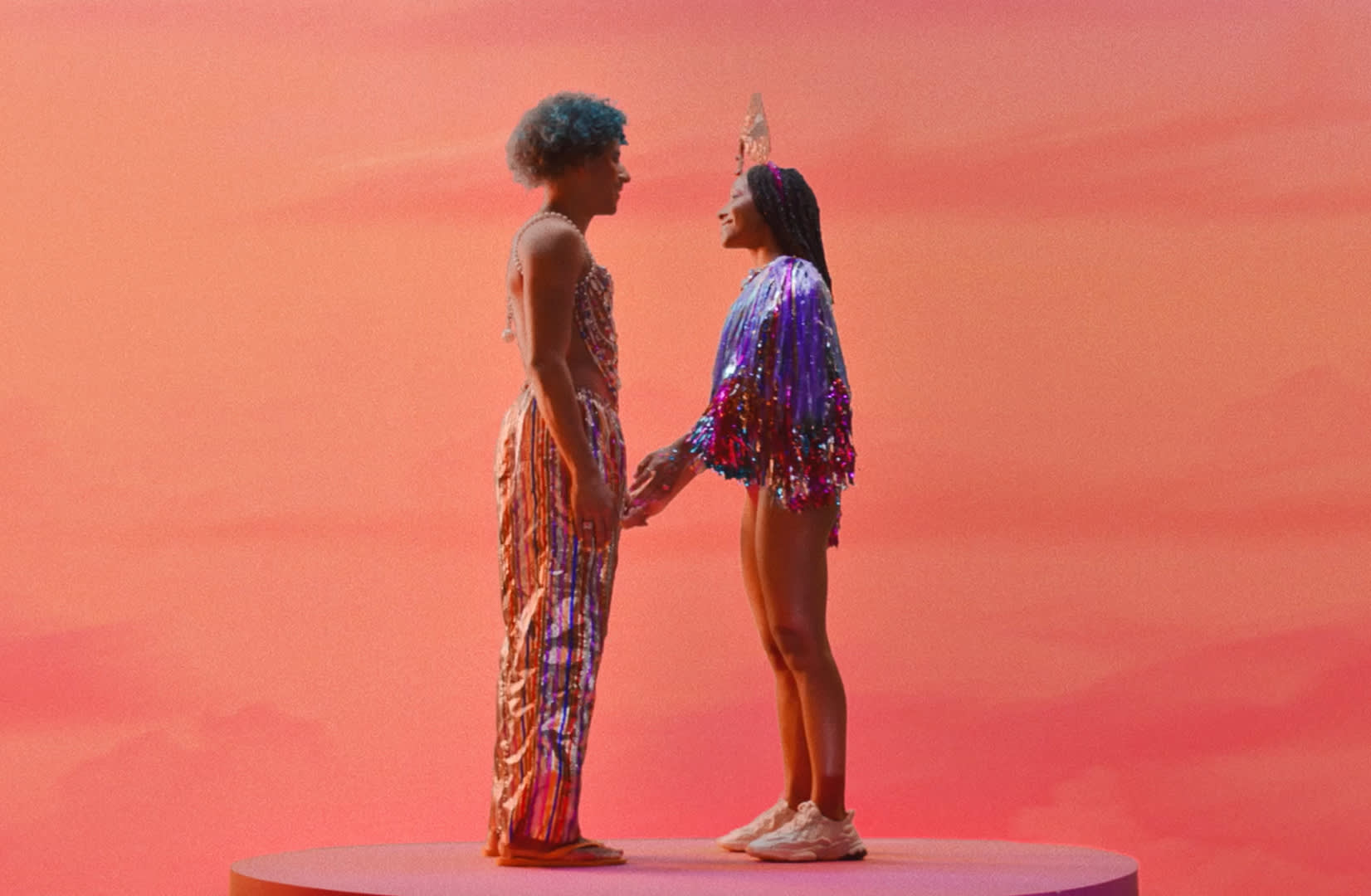 Two people stand facing each other on a round platform against a pink sunset backdrop. One wears a shiny, sequined outfit; the other wears a colorful, fringed top, shorts, sneakers, and a decorative crown. They hold hands and smile.