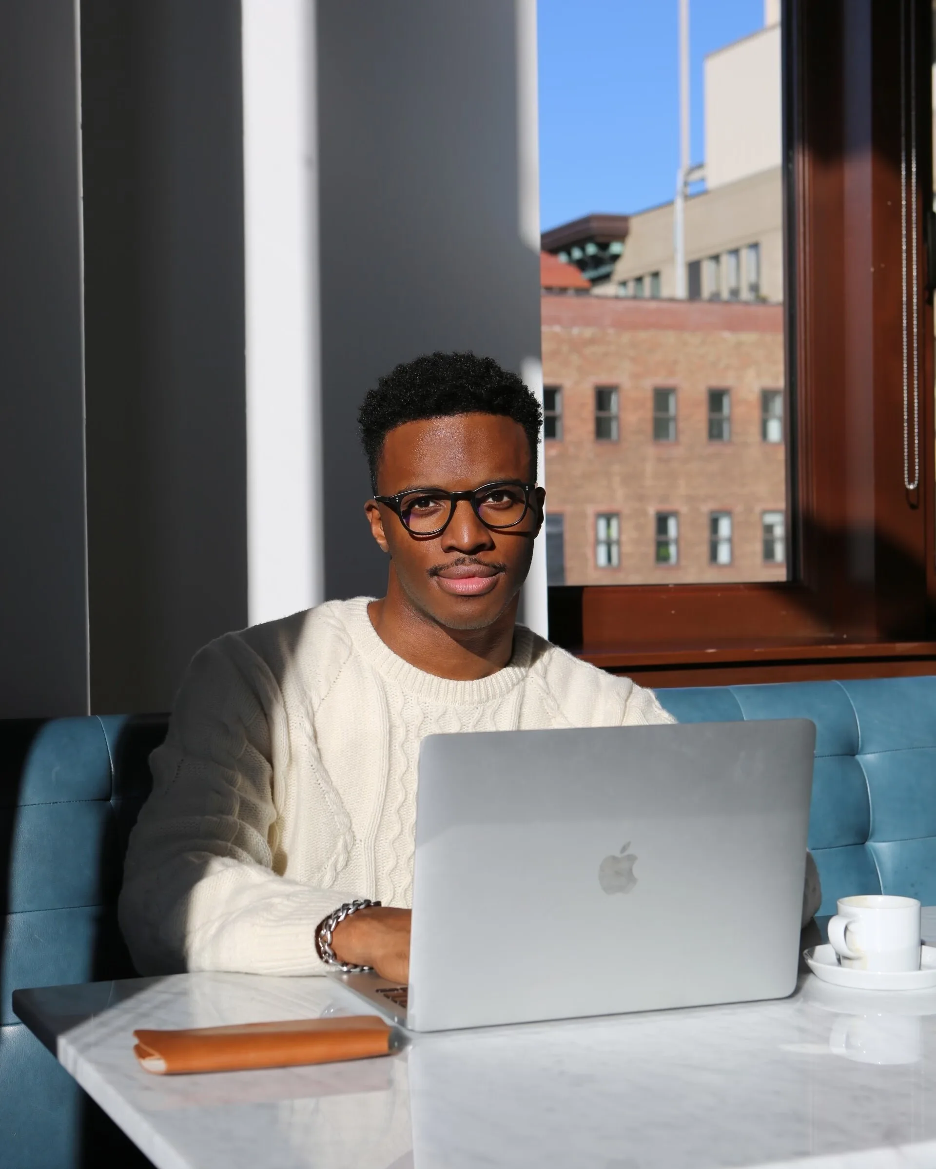 A person wearing glasses and a white sweater sits at a table with a laptop, notebook, and a cup, in a bright room with sunlight streaming through a window and a city building visible outside.
