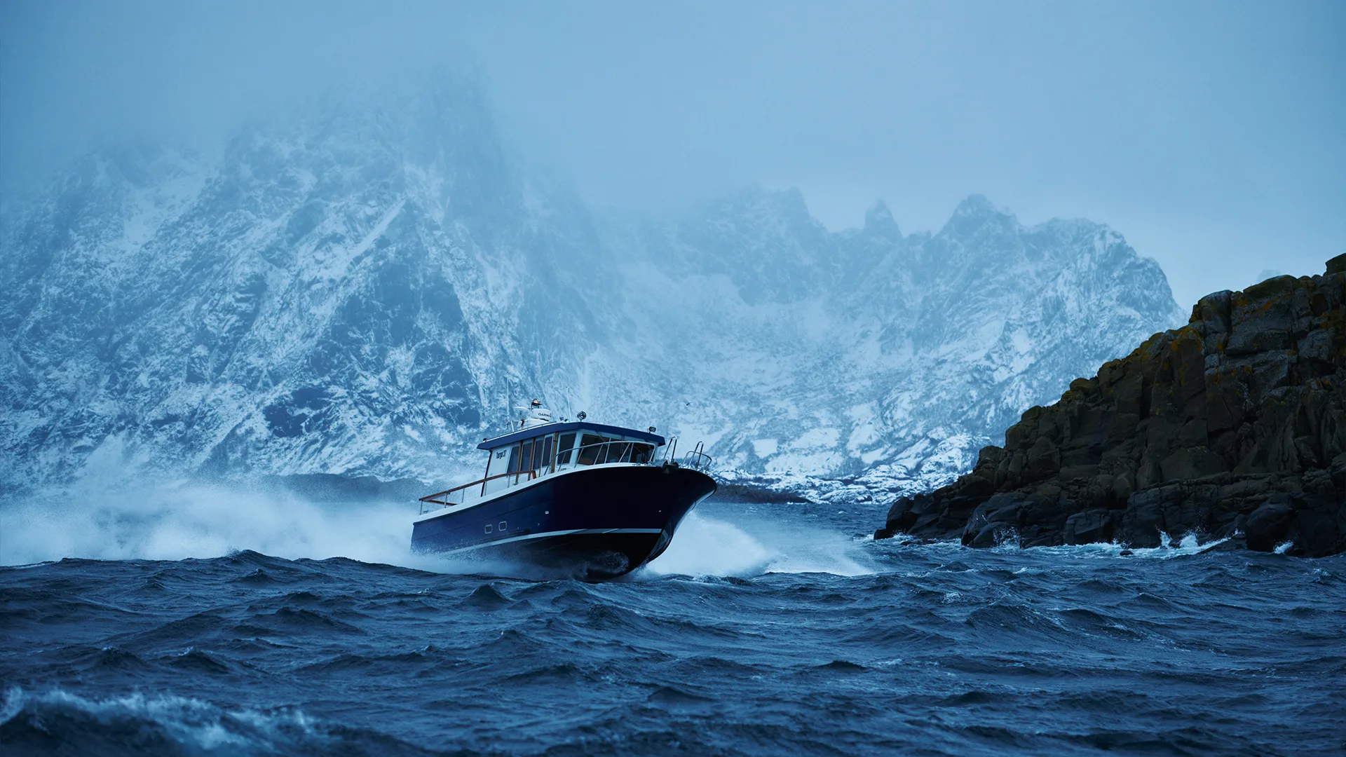 A boat speeds through choppy, dark blue water near rocky cliffs, with snow-covered mountains looming in the misty, cold background.