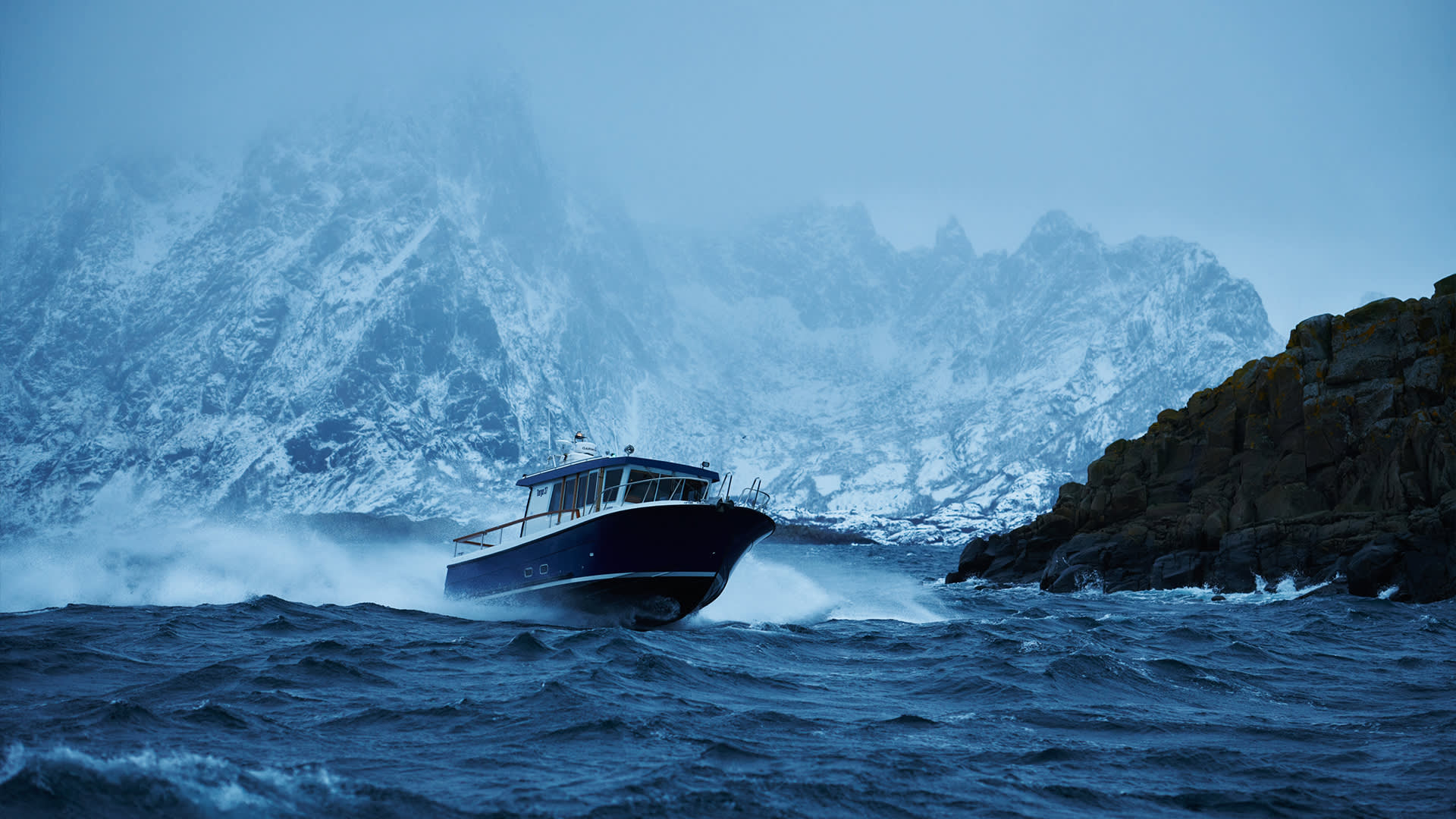 A boat speeds through choppy, dark blue water near rocky cliffs, with snow-covered mountains looming in the misty, cold background.