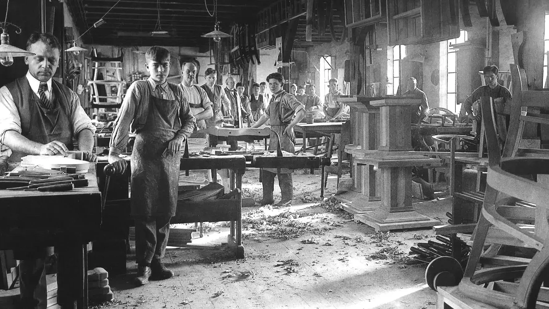 Black and white photo of men in aprons working in a carpentry workshop. Wooden tables, shavings on the floor, and unfinished furniture pieces fill the well-lit room with large windows.