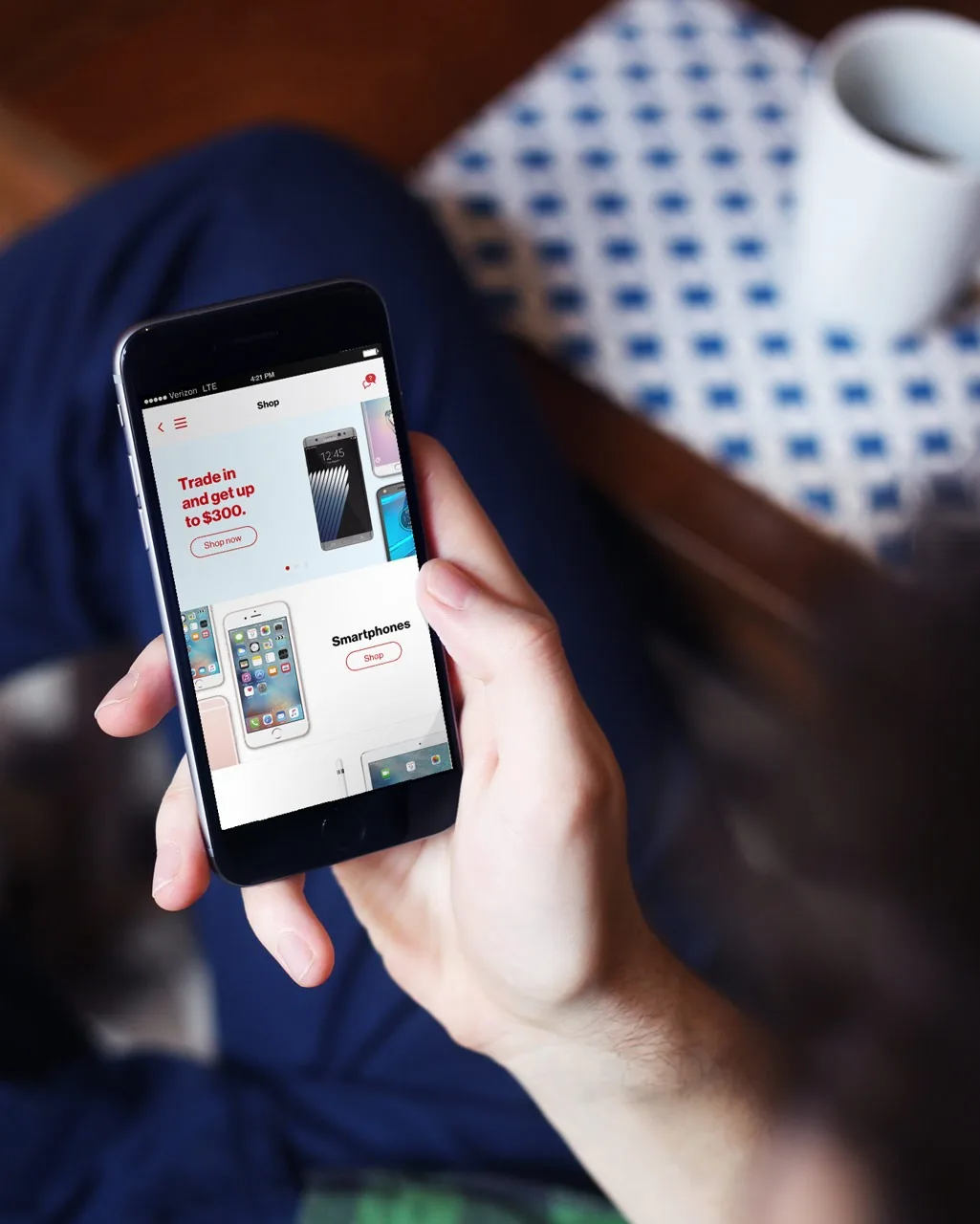 A person holds a smartphone displaying a shopping app, featuring offers for trading in devices for up to $300 and sections for smartphones. A white cup and a blue-patterned notebook sit on a table nearby.