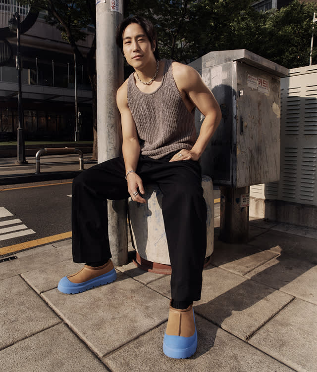 A man sits on a concrete post by a city sidewalk, wearing a gray sleeveless knit top, black pants, and tan shoes with bright blue soles. He gazes at the camera with trees and buildings in the background.