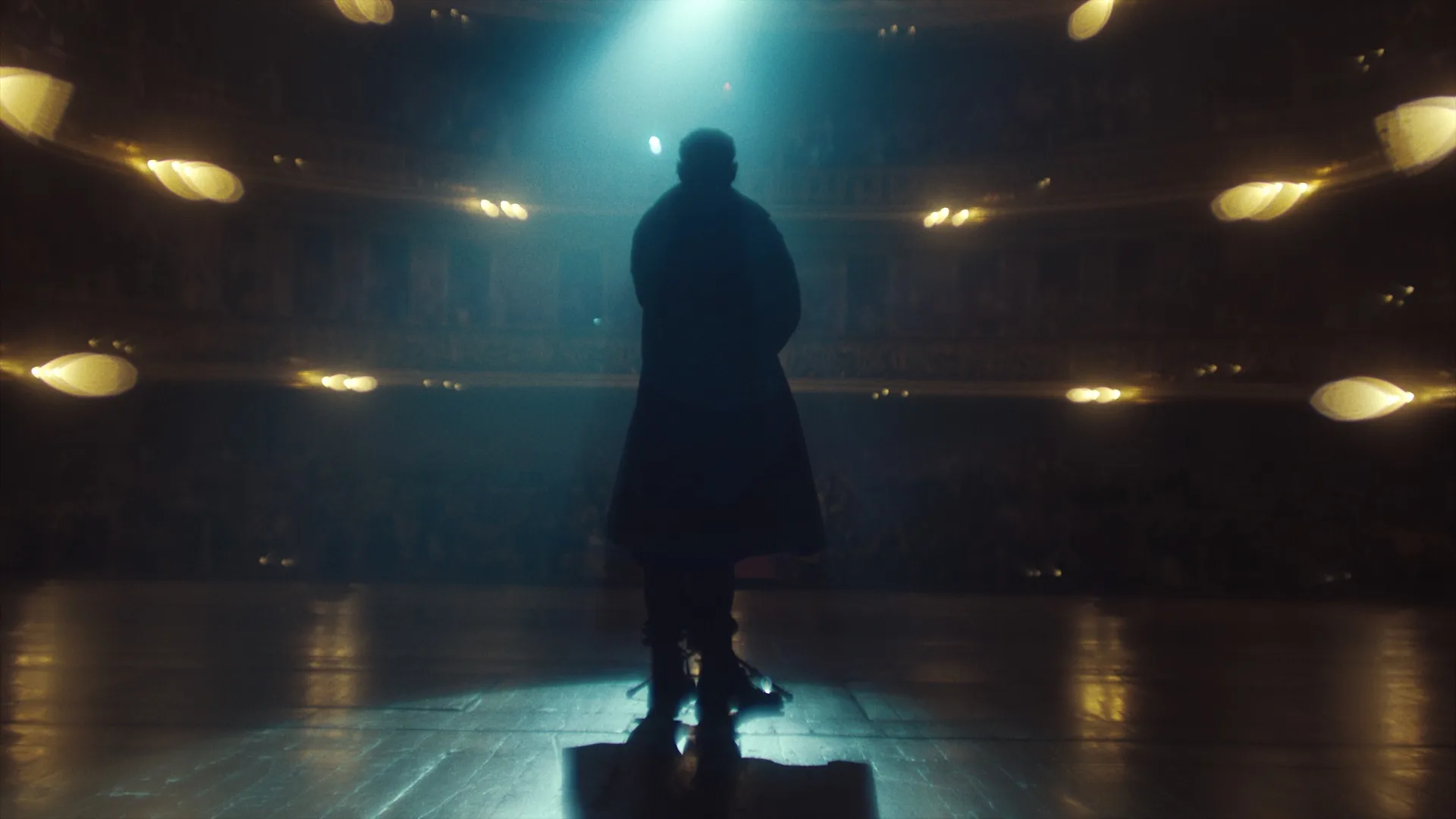 A person in a long coat stands alone on a theater stage under a bright spotlight, facing an audience seated in dimly lit, ornate balconies.