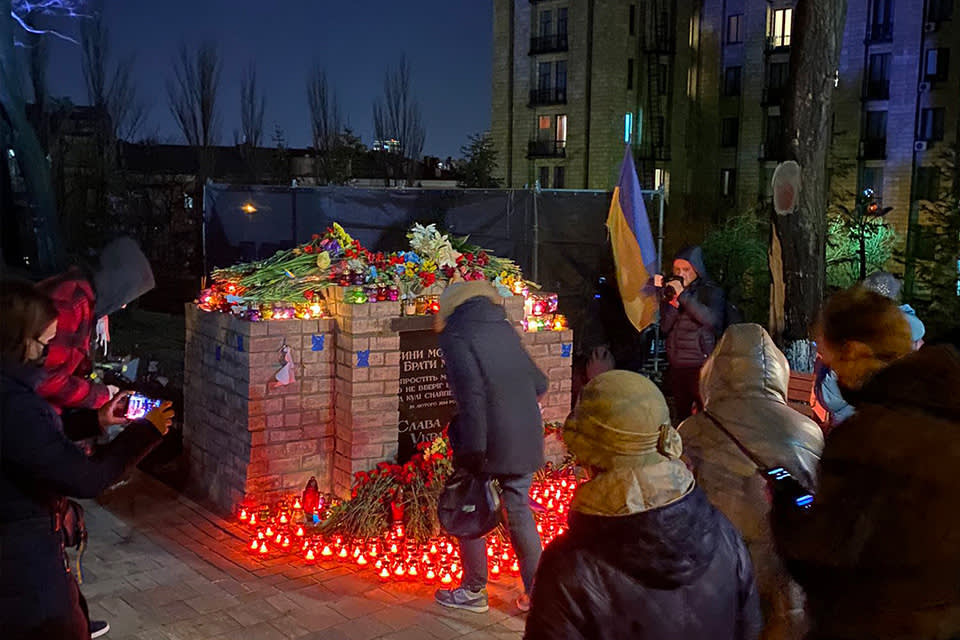 People gather at night around a memorial covered with flowers and lit candles. Some attendees hold Ukrainian flags and take photos, honoring and remembering victims. Tall buildings and bare trees are visible in the background.