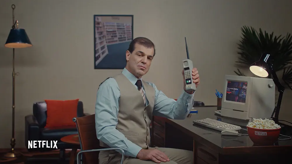 A man in a vest and tie sits at a desk holding a vintage cell phone. An old computer, popcorn, and a lamp are on the desk. A Netflix logo appears in the corner. The room has retro decor and a movie poster on the wall.
