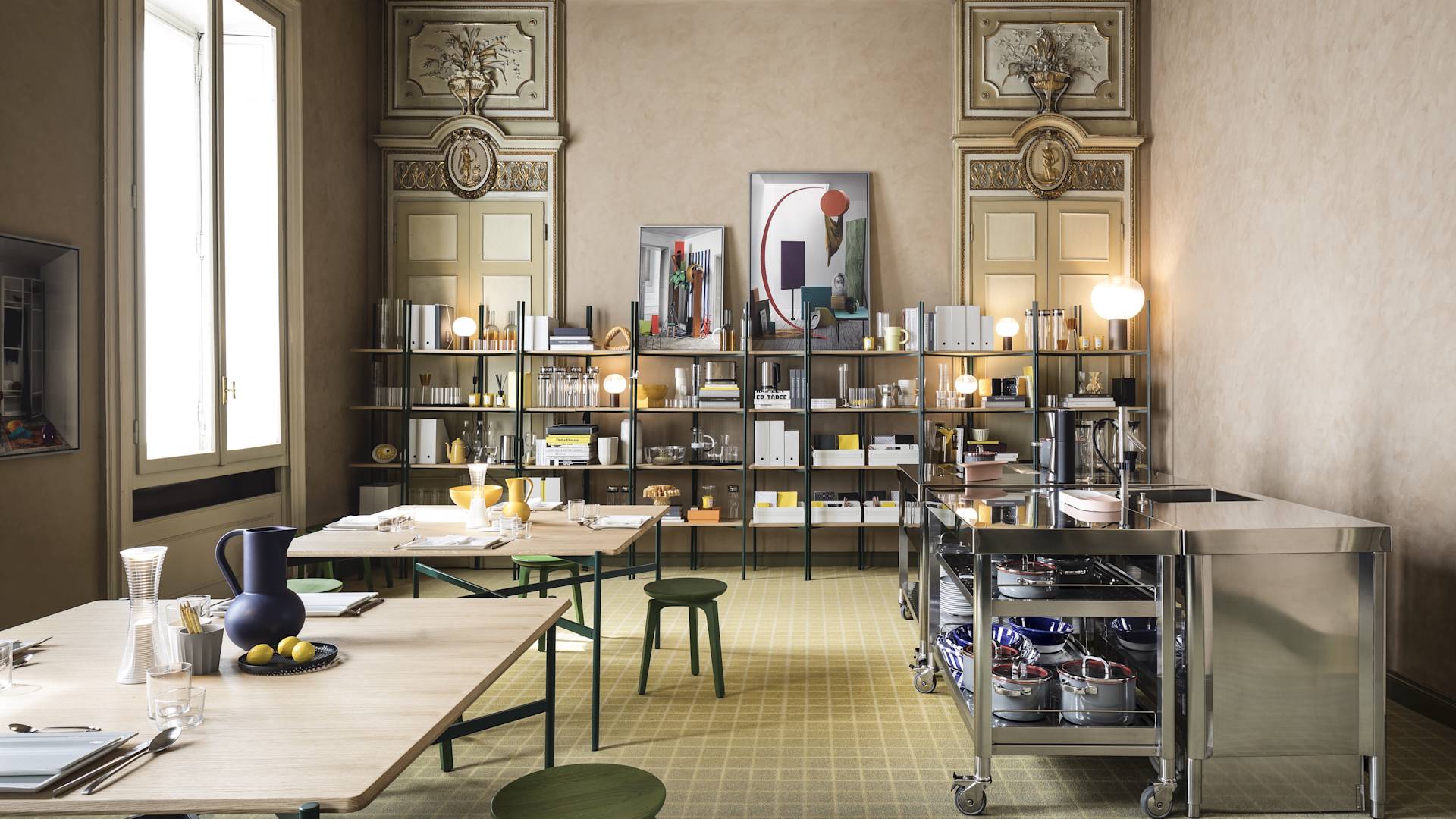 A modern, organized kitchen and dining space features shelves with books and decor, tables set with dishes, a metal cart with cookware, and large windows letting in natural light. Ornate wall details and a framed artwork add elegance.