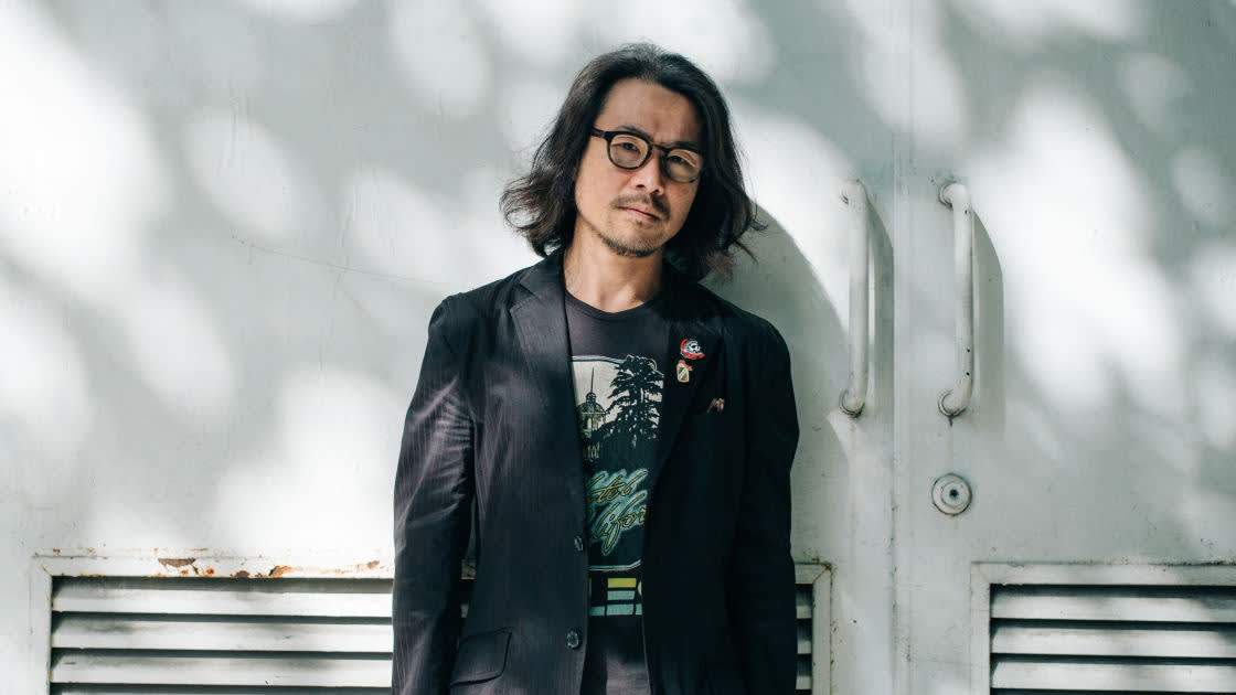 A man with shoulder-length hair and glasses, wearing a black jacket and graphic t-shirt, stands leaning against a white wall with double doors and vents, in dappled sunlight.